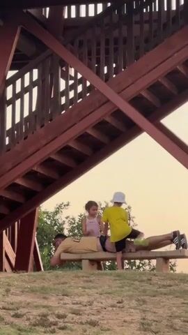 A man is lying on his stomach on a wooden bench under a wooden structure. Two children are standing on the bench behind him. The child on the left is a girl wearing a pink top. The child on the right is a boy wearing a yellow t-shirt and a white bucket hat. The boy is standing behind the girl, and they appear to be interacting with the man. The scene is outdoors with green grass and trees in the background. The sky is a pale orange, suggesting it is either sunrise or sunset.
