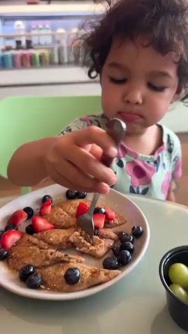 A child with curly brown hair wearing a floral print shirt is eating pancakes with strawberries and blueberries. She is using a fork to eat a piece of pancake. In the background, there are more pancakes and a cup of green grapes. In the foreground, there is a plate of food with potatoes, avocado, and what appears to be scrambled eggs or tofu scramble. The food is being eaten with a fork. There is also a glass of pink liquid and a cup of water on the table. The scene appears to be indoors, possibly in a restaurant.