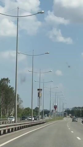 The video shows a view from a moving vehicle on a highway. There are streetlights with multiple lamps lining the road. There are also cars driving on the highway in both directions. On the right side of the road, there are several streetlights decorated with large, layered Russian flags. On the left side of the road, there are trees and a metal barrier. The sky is blue with some white clouds. The road is paved and has lane markings. The overall environment appears to be a sunny day in a semi-rural or suburban area.