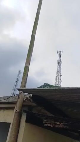 The video pans across a cloudy sky with a tall, thin telecommunications tower in the distance. In the foreground, a large green tree is visible, followed by a partial view of a dilapidated building with a corrugated metal roof and a concrete structure. The camera then moves to show more trees and vegetation, including some banana plants, against a backdrop of rolling hills obscured by mist or clouds. The overall scene suggests a rural or semi-rural environment under overcast weather conditions.