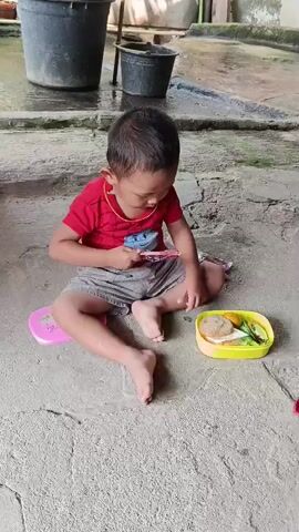 A young child wearing a red t-shirt and grey shorts sits cross-legged on a concrete ground. The child holds a pink object and appears to be eating it. To the child's right is a yellow and white lunchbox containing food. In the background, two black buckets and a building structure are visible. The ground is cracked and wet in some areas.