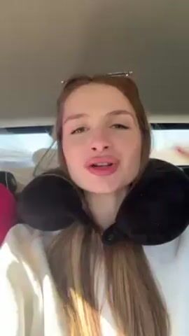 A young woman with long, light brown hair, wearing a white sweatshirt and a black travel pillow, is sitting in a vehicle and talking directly into the camera. She has sunglasses pushed up on her head and red nail polish on her fingers. She gestures with her hand as she speaks and lip-syncs along to music. The interior of the vehicle is visible behind her, including a gray headrest and some luggage. The scene appears to be filmed from the driver's seat or a similar position.
