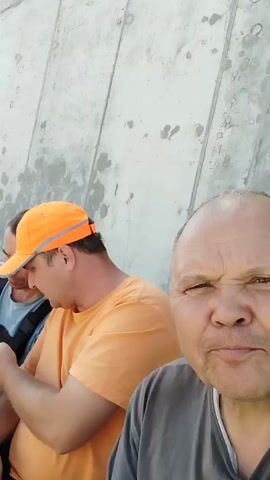 This short video appears to be a candid recording of a group of men, likely construction workers, taking a break outdoors. The subjects are positioned along a textured, light-colored concrete wall, suggesting they are on a construction site. The ground beneath them is covered in gravel.

Several men are visible, some seated and others in the foreground. One man in an orange baseball cap and an orange t-shirt is looking down at his phone. Another man beside him, wearing a blue t-shirt and an orange safety vest, is also engaged with his phone. A man in a grey bucket hat and dark work pants with reflective stripes is looking towards the camera, appearing somewhat serious. Further down the line, another man in an orange cap is partially visible. The man closest to the camera, who is likely holding the camera or phone, is looking directly into the lens with a pensive or perhaps slightly strained expression. His face is close to the camera, showing detailed textures.

The activity appears to be a moment of rest during a workday. The men are casually dressed in work attire, including reflective elements on their pants and safety vests. One man is holding what appears to be a black object, possibly a tool or a piece of equipment.

The lighting suggests it is daytime, likely late morning or afternoon, with a bright but possibly overcast sky, given the lack of harsh shadows. The concrete wall has areas of staining and wear, indicating it's an active work environment. There is no visible text in the video.