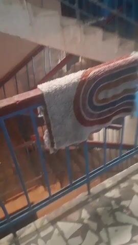 The video shows a section of an indoor staircase with a blue and red railing. A rug is draped over the railing. The walls are painted a light color and have some discoloration. The floor appears to be tiled with broken pieces. The video pans from a close-up of the rug to a wider view of the staircase and then to another section of the landing with a blue wall and some items on the floor.
