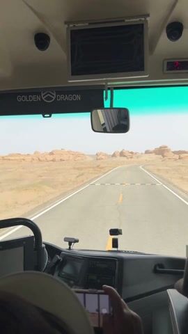 A point-of-view video from inside a "GOLDEN DRAGON" brand bus shows a desert landscape through the front windshield. The bus is driving on a paved road that curves slightly to the right, with a yellow dividing line and white shoulder lines. On either side of the road, sandy desert stretches out, dotted with distinctive, eroded rock formations known as yardangs. The sky is bright and appears slightly hazy or dusty.

Inside the bus, the dashboard, steering wheel, and a rearview mirror are visible. An overhead monitor and a digital display showing "2:0" and then "0" are also present. The hands of a person are visible in the foreground, holding a smartphone horizontally and filming the view ahead. The scene suggests a journey through a scenic, arid region, likely in Dunhuang City, China, known for its desert landscapes and yardang formations. The time of day is daytime, with clear visibility of the distant rock formations.