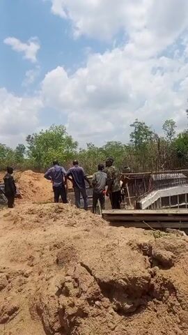 The video depicts four men standing near a construction site, observing a concrete structure with reinforcing steel. The surrounding environment consists of sandy soil, trees, and a cloudy sky. A black water tank is visible near a pile of dirt. One man walks away from the construction site, carrying what appears to be a tool.