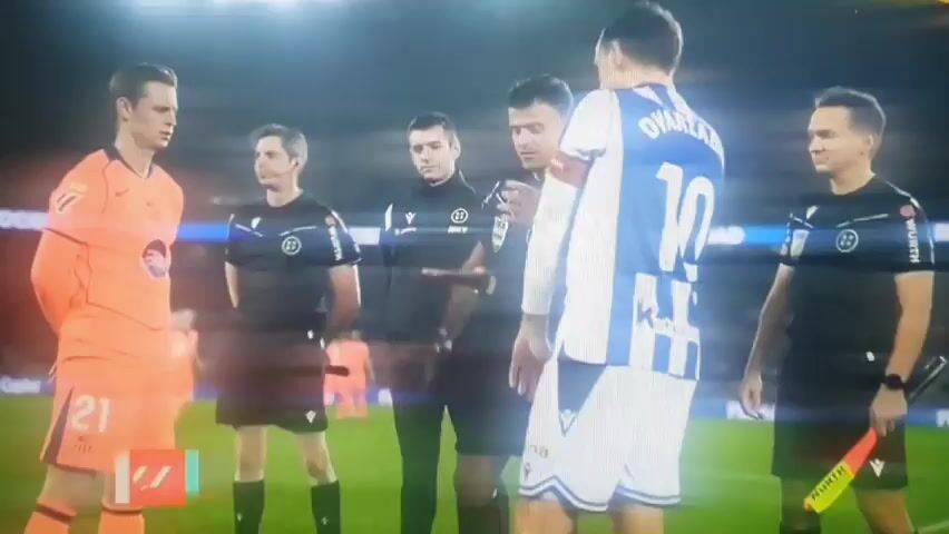 Five people stand on a soccer field, with four wearing referee uniforms, and one wearing a blue and white soccer uniform with the number 10. The referee uniforms are black, with blue accents and a FIFA patch. One referee wears an orange uniform. They are facing each other and gesturing. The camera focuses on the center referee who raises his hands and appears to be talking. A graphic on the left side of the screen lists the referees: Jesús Gil as referee, Ángel Nevado and Javier Martínez as assistants, and Daniel Miranda as fourth referee. The ball of the match is listed as LA LIGA ORBITA YELLOW. The environment is a brightly lit soccer stadium.