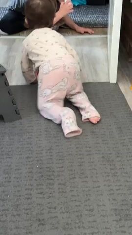 The video shows a young child, likely a toddler, crawling on a gray carpet. The child is wearing pink pajamas with a bunny pattern. The child is moving towards a doorway. In the background, another person is visible, possibly an adult, partially obscured by the doorway and the child. The environment appears to be indoors, possibly a home, with a step stool visible to the left of the frame. The lighting is moderate.