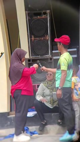 A woman wearing a pink hijab and a pink shirt is speaking into a microphone. She is holding a piece of paper in her left hand. She is standing on steps next to a large speaker. Another woman wearing a green hijab and sunglasses is sitting on the steps next to the speaker. A young boy is standing next to the sitting woman. A man wearing a red hat and a green and yellow t-shirt is standing to the right of the woman with the microphone, holding the microphone. The environment appears to be outdoors, possibly at an event.