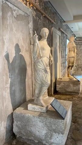 The video pans across an indoor exhibit featuring ancient marble statues displayed on rough-hewn stone bases. In the foreground, a white marble statue of a draped female figure stands, her left arm raised, holding a slender object. Her gaze is directed forward. To the left of this statue, a distinct shadow of a similar figure is cast on a textured wall. Further back and to the right, another white marble statue of a male figure in a toga-like garment is visible. The walls appear to be constructed of stone and plaster, with wooden beams supporting the ceiling. A dark informational plaque is positioned in front of the female statue. The floor is paved with cobblestones. The lighting is dim, suggesting an indoor museum or historical site.