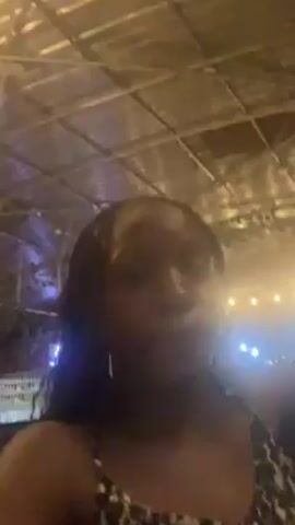 The video shows a Black woman standing indoors. She is wearing a black and white patterned top. The woman is looking at the camera and talking, sometimes looking up and to the side. Her hair is styled in braids, and her face is lit by ambient light. Overhead, a metal grid-like ceiling is visible, with various lights hanging in the background. The video seems to be taken at night, as the lighting is dim, and there appear to be other people and objects in the background, though they are out of focus.