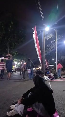 A group of people are gathered outdoors at night. In the foreground, a person wearing a black hoodie and white shoes is sitting on a pink bag. Further back, several other individuals are standing and sitting on the ground, some near a food stall with a blue tarp. A motorcycle is parked on the side of the road. A tall, multi-story building is visible behind the gathering. A red and white flag is draped over a pole, illuminated by a street light. The overall environment appears to be a street or public area.