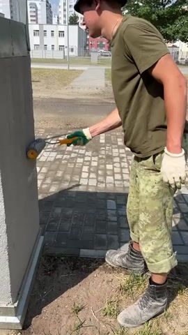 A young man wearing a green t-shirt, camouflage pants, a blue bucket hat, and work boots is standing outside and using a paint roller to apply a light-colored substance to the side of a grey, textured structure. He is also wearing white gloves. The structure appears to be a small, free-standing building or utility box. The ground is covered with grey pavers and some dirt and grass. In the background, there are apartment buildings, trees, and a paved area. The sun is shining, and the lighting suggests it is daytime.