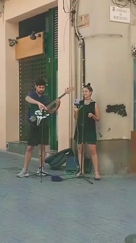 Two musicians perform on a pedestrian street corner. A man stands on the left playing a bass guitar, while a woman stands on the right singing into a smartphone attached to a microphone stand. The woman is dressed in a black dress and sandals, and her hair is tied in a bun. The man wears a dark blue t-shirt, black shorts, and sneakers. Their equipment includes two microphone stands, a small amplifier, a bass guitar case and a purple object, likely a pedal, on the ground. The corner building has a closed storefront with a green-painted frame on the left and a green-painted door on the right.  A street sign above reads "Avinguda de Barcelona". The street is paved with gray tiles.