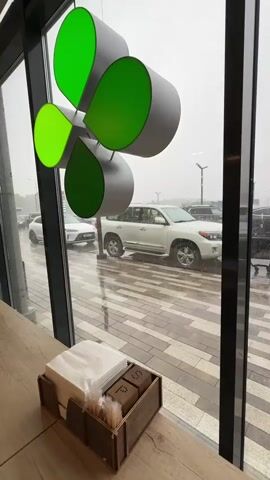 The video shows a view through a large window into a parking lot on a rainy day. In the foreground, on a wooden table, is a wooden caddy holding napkins and salt and pepper shakers. The caddy is made of dark wood with visible grain and has labels on the shakers that appear to read "S" and "P". Above the table and caddy, a decorative mobile hangs from the ceiling. This mobile consists of four grey, curved, petal-like shapes with bright green interiors, arranged in a cluster.

Through the window, the parking lot is wet and reflects the grey, overcast sky. Several vehicles are visible, including a white SUV, a silver SUV, and other cars. Some vehicles appear to be moving, while others are parked. A black shopping cart is also present in the parking lot, with someone pushing it. The ground of the parking lot is paved with rectangular tiles in a pattern of light and dark grey. The overall impression is of a dreary, wet day, viewed from indoors.