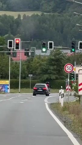 A black SUV and a motorcycle are stopped at a traffic light. The traffic light for the SUV is red, and the traffic light for the motorcycle is green. There is a speed limit sign of 50. The environment appears to be a rural road with trees and hills in the background. The sky is overcast.