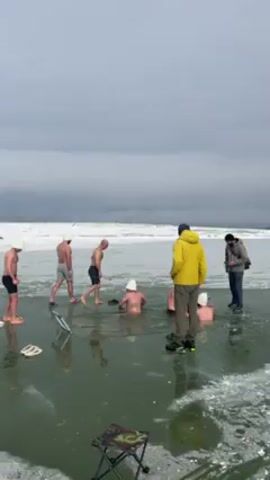 The scene captures a group of individuals engaging in what appears to be an ice bath or winter swimming event. Six men are partially submerged in icy water, and some are wearing white hats. Two men, fully clothed, stand on the edge of the ice, seemingly observing or supervising. One is in a yellow jacket, and the other is in a darker coat. The environment is predominantly icy and snowy, with a grey sky overhead. A foldable chair and a pair of flip-flops lie on the ice. One of the men is entering the water. The men in the water are interacting with each other. The atmosphere seems cold and challenging.