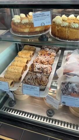 The video pans across a bakery display case filled with various desserts. On the top shelf, two cheesecakes are visible, one labeled "Apple Caramel Crumble Cheesecake" priced at 9.50. To the right, several servings of what appears to be tiramisu are stacked. On the middle shelf, a row of sausage rolls is on the left, followed by what looks like croissants or pastries with a drizzled topping, then cherry clafoutis priced at 6.75. Further to the right are rows of chocolate cakes topped with pecans, labeled "Espresso Pecan Chocolate Cake." Next to those are rows of purple-glazed eclairs, labeled "London Fog Éclair" priced at 7.50. To the right of the eclairs are lemon meringue tarts priced at 8.50, and strawberry tarts, labeled "Fresh Fruit Tart" priced at 8.50. The bottom shelf has orange juice bottles. The display case is well-lit and appears to be inside a clean, modern establishment.