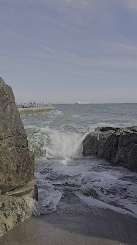 The video displays a dynamic ocean scene with waves crashing against rocky outcrops in the foreground. In the mid-ground, a man-made structure, possibly a pier or a walkway with railings and several people on it, juts out into the water. Beyond this, a large ship is visible on the horizon, sailing across the vast expanse of the sea. The sky is clear with a few wispy clouds, suggesting fair weather. The lighting indicates it is daytime, likely mid-morning or afternoon, given the brightness and shadows. The overall mood is one of natural power and tranquility, with the crashing waves providing a sense of energy against the calm, expansive seascape and the distant vessel. There is no visible text in the media file.