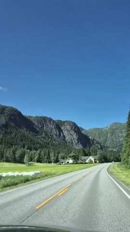 This is a first-person perspective video taken from a vehicle driving on a paved road. The road has a yellow double line in the center and white lines on the sides. The surrounding environment is mountainous and rural. On the left side of the road, there is a large green field with some white cylindrical objects, likely hay bales, and a cluster of buildings that appear to be houses or farm structures. Behind the field and buildings, a steep, rocky mountain covered in dense green forest rises sharply. The sky is a clear, bright blue. On the right side of the road, there is a green grassy verge with wildflowers and more forest. The video shows the road curving gently to the right as the vehicle continues to move forward. The overall impression is of a scenic drive through a natural landscape on a sunny day.