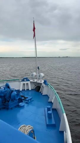 The video shows a view from the bow of a boat moving on a body of water. The deck of the boat is blue and has some equipment on it, including a rope and a winch. A flag with a red design is flying from a pole. The water is calm with small waves. In the distance, there is a line of land with some trees. There is also a small inflatable boat with at least two people in it visible in the distance, moving across the water. The sky is overcast with clouds.