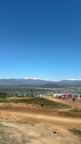 The video shows a motocross course on a sunny day with a clear blue sky and distant mountains capped with snow. Several motorcyclists are visible on the course, performing jumps and navigating the dirt terrain. Spectators are gathered near the course, standing behind fences and under tents. A person wearing a black and white outfit is walking alongside the track. The environment consists of a dry, dusty landscape with a small town visible in the distance.