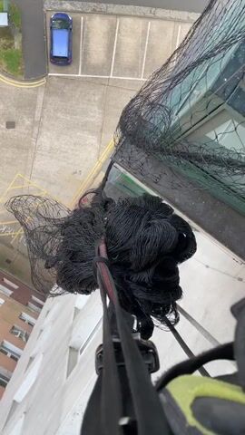 The video captures the installation or maintenance of a large black bird control net on a high-rise building in Hertsmere, United Kingdom, during daytime with clear weather.

The initial shot provides a first-person, high-angle view looking down from the building. A substantial black net, bunched up and attached to a rope or strap, is being handled by an unseen person. Below, a street, a car park with a blue car, and other buildings are visible. Faint yellow markings are on the ground, but any text is indistinct.

The next sequence shows the net partially unrolled, covering a section of the building's exterior. A worker's foot is briefly visible at the top right, suggesting they are positioned on a ledge or roof. Further close-up shots focus on the net's installation details, showing it secured to a textured, light-colored concrete or stucco wall with metal fasteners. The net is then seen covering a dark green window frame, indicating its purpose to prevent birds from accessing or nesting on the building's surfaces. No discernible text is clearly visible throughout the video.