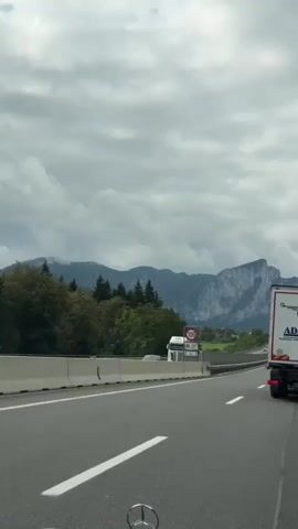 The video begins with a shot of a highway with a white convertible car driving in the right lane. Mountains are visible in the background. A truck is driving in the left lane. A speed limit sign showing 100 is visible on the side of the road. The camera is positioned inside another vehicle, showing part of its dashboard. The sky is cloudy. The convertible car passes the speed limit sign and continues down the highway. The truck remains in the left lane. The scenery of the mountains and trees remains in the background. The video ends with the white convertible car continuing to drive down the highway.