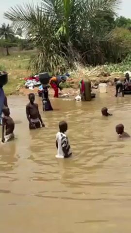 The video shows a group of children playing in a muddy, brown-colored body of water. Some of the children are standing, while others are swimming or splashing each other. On the shore, several women are doing laundry in tubs. Some of the women are carrying buckets on their heads. The background includes a palm tree, other vegetation, and what appears to be discarded items on the bank.