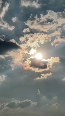 The video shows a cloudy sky with the sun shining through the clouds. The sun is bright and appears as a white orb in the center of the frame. The clouds are a mixture of dark and light gray, with some areas illuminated by the sun. There are rays of sunlight visible, creating a dramatic effect. The overall mood is atmospheric and peaceful. The video is shot from a low angle, looking up at the sky.