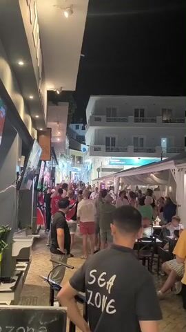 The video captures a crowded street scene at night, possibly a resort town. The narrow street is lined with buildings, some with balconies, and open-air establishments with white canopies that appear to be restaurants or bars. Numerous people are walking along the street, mingling, and standing in groups. Many are dressed casually in summer attire. Some individuals are holding up their phones, presumably to take pictures or record videos. The atmosphere appears lively and social. Flags, possibly national flags, are visible hanging on a building on the left side of the street. Artificial lighting illuminates the scene, creating a festive evening ambiance.