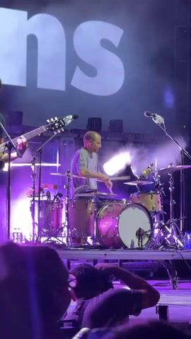This is a vertical video clip of a live music performance, likely at a concert. The central subject is a bald male drummer seated behind a drum kit. He is wearing a grey t-shirt and appears to be actively playing the drums, holding drumsticks in his hands. The drum kit is prominently featured, with several drums and cymbals, all appearing to have a gold glitter finish.

To the left of the drummer, partially visible, is a person playing an electric guitar. The guitar is dark-colored. To the right of the drummer, another person's arm and shoulder are visible, suggesting another band member.

The stage is lit with vibrant purple and bright white lights, creating a dramatic atmosphere. Smoke or haze is visible on stage, particularly around the lighting. In the background, a large white sign with the letters "ns" is displayed prominently on a dark surface, possibly a backdrop or screen. The overall setting appears to be an indoor venue or an outdoor stage at night, given the artificial lighting and the darkness surrounding the stage.

In the foreground, the blurry silhouettes of concertgoers are visible, with some of their heads and shoulders in focus. The perspective is from within the audience, looking up at the stage. The mood conveyed is energetic and exciting, typical of a live concert performance.

No specific time of day or weather conditions are discernible, but the event is clearly taking place in Kragujevac, Serbia.