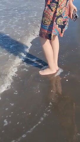 A person stands on a wet sandy beach with waves gently lapping at their bare feet. The person is wearing a colorful patterned dress and holding a smartphone. The sun casts a shadow on the sand.