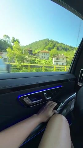 The video shows the interior of a luxury car, likely a Mercedes-Benz, with a person's hand operating the window controls. The window shade is being lowered, revealing a scenic view of houses and trees on a hillside under a clear blue sky. The interior of the car features ambient blue lighting, a leather door panel, and a speaker grille. The focus is on the window controls and the changing view outside as the shade is adjusted.
