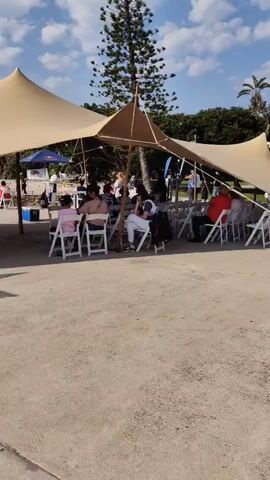 This media file captures an outdoor event taking place under a large, tan stretch tent. Several people are seated on white folding chairs, facing away from the camera towards an unseen focal point. The tent provides shade from a bright, sunny day with a partly cloudy sky. In the background, lush green trees and palm trees suggest a warm climate.

People are dressed casually, with a mix of adults and children present. The atmosphere appears relaxed, with individuals engaged in conversation or observing something in front of them. A young girl in a pink dress is visible in the foreground, seated in a white chair. Further back, other attendees are enjoying the event.

Notable objects include a tall, thin tree behind the tent, which could be a pine, and what appears to be a Red Bull umbrella in the background. There are also some signs visible, one of which has the Red Bull logo. The ground is a light-colored, paved surface. The scene suggests a community gathering, possibly a festival, market, or performance, taking place in Gqeberha, South Africa. The time of day is likely afternoon, given the bright sunlight and the shadows cast by the tent poles.
