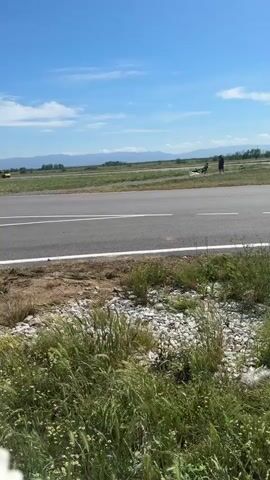 The video shows an airport runway on a sunny day. The camera is positioned low to the ground, with grass and weeds in the foreground. In the distance, mountains can be seen. A yellow vehicle is parked far down the runway on the left. A race car speeds across the runway. After it passes, a group of people are standing near a damaged vehicle on the right.
