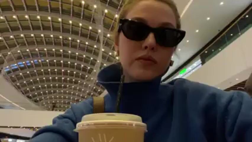 A woman wearing sunglasses and a blue sweater is drinking from a takeout cup with a straw in it. The cup has a simple white sun-like design. She is indoors in what appears to be a shopping mall. The ceiling is high with a modern grid pattern and small lights. She takes a sip and looks directly at the camera.