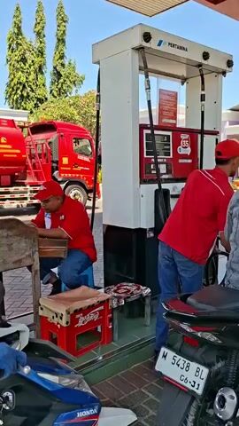 At a Pertamina gas station, a male attendant in a red shirt and blue jeans stands near a gas pump, holding what appears to be a payment device. Another attendant, also in a red shirt and blue jeans, is seated to the left, looking down at a wooden surface. In the foreground, parts of two motorcycles are visible, one white with blue and red accents, and another mostly black. Behind the seated attendant, a red fuel tanker truck is parked. The environment is outdoors, under a covered area, with trees and buildings visible in the background under a clear sky.