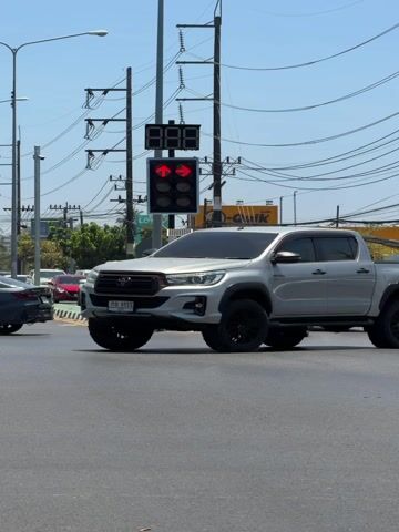 The video captures a sunny daytime scene at a busy intersection. The sky is clear and blue, suggesting good weather. In the foreground, a gray pickup truck with black wheels is partially visible on the left, moving towards the right side of the frame. A light gray electric car (possibly a NETA V) with distinctive front lighting is seen turning left across the intersection, followed by a black sedan and a white sedan also making a left turn. Several other cars, mostly sedans in various colors like white and red, are visible further back, either waiting at the traffic light or also turning. A person on a light blue scooter is also visible, briefly in the first few seconds, appearing to move alongside the turning cars.

A prominent traffic light pole stands in the center of the frame. It features two sets of traffic signals: one displaying a red countdown timer showing "888" (likely a placeholder or malfunctioning display, as it doesn't count down sequentially in the clip), and another below it with two red arrows pointing left and right, indicating that vehicles in those lanes should stop or yield. A blue circular sign with a white arrow pointing left is attached to the pole, indicating a left turn.

Numerous power lines crisscross the upper part of the frame, strung between utility poles. In the background, on the right side, there's a large yellow building or container with "Q-quick" text visible, along with some green trees. The road surface is asphalt. License plates are visible on some of the vehicles: the gray electric car has "ขท 6371" and a white sedan later in the clip has "ฎก 8423" and another white car has "ณข 5828". The overall impression is a typical urban or suburban intersection in Southeast Asia, given the vehicle types and general infrastructure.