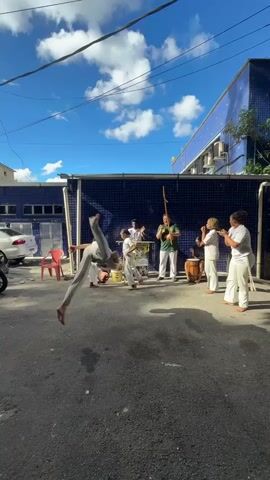 A medium, outdoor, daylight shot shows a capoeira group with nine people on a paved street. The street is partially shaded, and bright sunlight is visible on the street and on the buildings in the background. The street is paved in asphalt. A building to the left, which has a partially visible white car parked in front of it, is blue with white horizontal stripes and a large window. A building to the right, which also has a partially visible white car parked in front of it, is white. A person in the foreground, wearing a white shirt and grey pants, is doing a handstand, with their legs in the air and their head near the ground. Another person, wearing a white shirt and white pants, is standing behind them and looking at the camera. A person in the mid-ground, wearing a green shirt and white pants, is holding a berimbau. Another person, wearing a white shirt and white pants, is standing behind them and playing a drum. Other people are in the background, some wearing white shirts and white pants, and some wearing white shirts and white pants and doing capoeira moves.