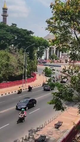 The video shows a street scene from an elevated perspective on a sunny day. The road is paved and has multiple lanes. On the left side of the road, there is a red barrier with white poles. Beyond the barrier is a sidewalk with benches and trees. On the right side of the road, there are trees and a building with large columns. In the background, a minaret of a mosque is visible. Vehicles, including cars and motorcycles, are moving on the road. Several people are riding motorcycles. One car has yellow markings on its side. The sky is bright and appears to have some clouds.