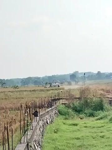 A white helicopter lifts off from a field, kicking up dust. The field is bordered by a wall with wooden stakes on one side and green grass on the other. Trees and buildings are visible in the background.