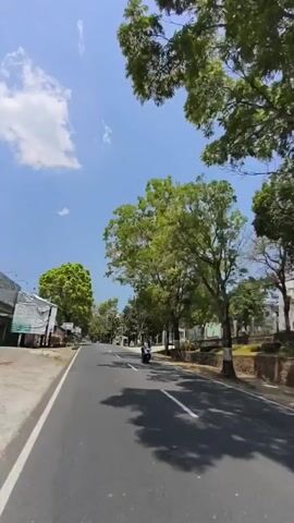 A motorcycle with two people on it rides down a paved road lined with trees and buildings. The sky is blue with some white clouds. The motorcycle is in the right lane, and there is a white line in the middle of the road. There are buildings on both sides of the road, and some of the buildings have awnings. There are also some trees on both sides of the road. The motorcycle is moving away from the camera.
