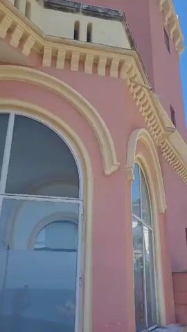The video shows a coastal scene, starting with a view of a pink building with arched windows and decorative moldings against a clear blue sky. The camera pans to reveal a beach with white sand, a few people sunbathing, and a white parasol. Buildings are seen on a hill overlooking the beach. The camera continues to pan, showcasing the clear, turquoise water of the sea, with some people swimming and relaxing on inflatable tubes. A stone wall separates a small beach area from the water. Green vegetation covers the top of the coastal cliffs.