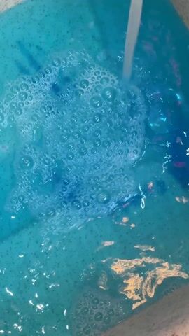 The video depicts a container being filled with water, which is creating numerous bubbles. The water has a bright blue hue and contains some small particles, giving it a slightly textured appearance. There also appears to be some iridescent material or glitter scattered throughout the water.