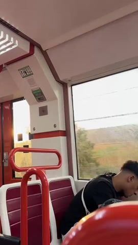 The video shows the interior of a train. The seats are red and white, with red grab bars. A young man in a black t-shirt and white crossbody bag is visible through the window, appearing to be asleep or resting his head on his arms. The scenery outside the window is blurred, suggesting the train is in motion, and consists of fields and trees under a bright sky. The lighting appears to be natural, possibly from the setting sun, casting a warm glow. The overall impression is a quiet moment during a train journey.
