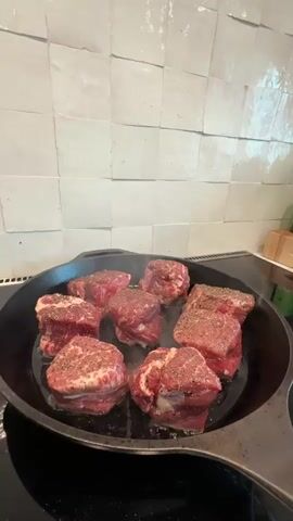 This is a close-up shot from a high angle, looking down at a black cast iron skillet filled with eight cuts of seasoned raw beef. The steaks are arranged in a rough circle within the pan, with one larger rectangular cut on the right side. They are seasoned with salt and pepper and are beginning to sear, as evidenced by the visible steam rising from the pan. The skillet is placed on a black stovetop with a white tiled backsplash in the background. The lighting is bright, illuminating the details of the meat and the cooking surface. The video appears to be a short, looping clip, capturing the initial stages of cooking the steaks.