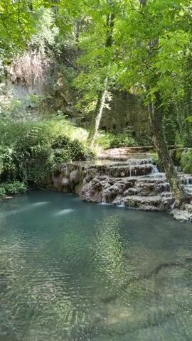 The video shows a serene natural scene with a waterfall flowing over a series of rocky tiers into a clear, turquoise pool. Lush green trees and vegetation surround the waterfall, with sunlight dappling through the leaves. The water appears calm and inviting, with ripples on its surface. The overall atmosphere is peaceful and tranquil, evoking a sense of untouched nature. There are no people or man-made objects visible in the video, only the raw beauty of the natural environment. The camera appears to be stationary, capturing the continuous flow of water and the play of light and shadow. The video captures the essence of a secluded natural oasis, a place of beauty and tranquility.