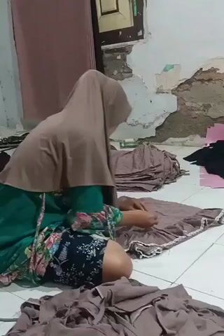 A person wearing a hijab and a green dress with a floral pattern over dark patterned shorts is kneeling on a tiled floor. They are working on a piece of brown fabric with white lace trim, and there are piles of similar brown fabric scattered around them. The environment appears to be a room with damaged walls and a pink curtain in the background.