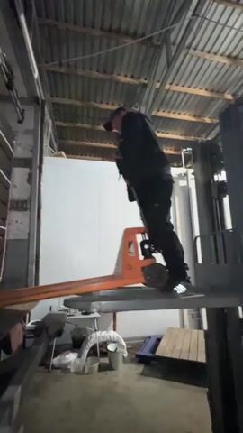 A man in a black hoodie and dark pants, wearing a baseball cap and reflective-striped shoes, operates a manual pallet jack. He is standing on a raised platform within what appears to be a warehouse or industrial setting. The pallet jack is orange and has its forks extended, supporting the platform he is standing on. He is looking down and appears to be maneuvering the pallet jack. In the background, there are shelves filled with items, possibly boxes or crates, and a large white wall. The ceiling has exposed beams and corrugated metal. The floor is concrete. To the left, there is another piece of equipment, and to the right, there are wooden pallets. The lighting is dim, suggesting an indoor, possibly evening or early morning, environment.